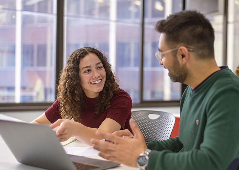 A man and a woman sit at a table. They are talking and smiling. A laptop is on the desk. 