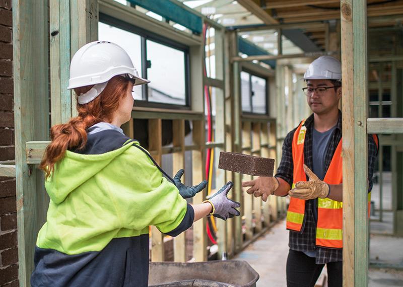 Two people working on a construction site with one passing the other a brick