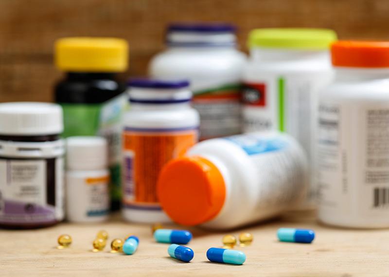 Several pills lie on a table with an assortment of pill bottles in the background. 