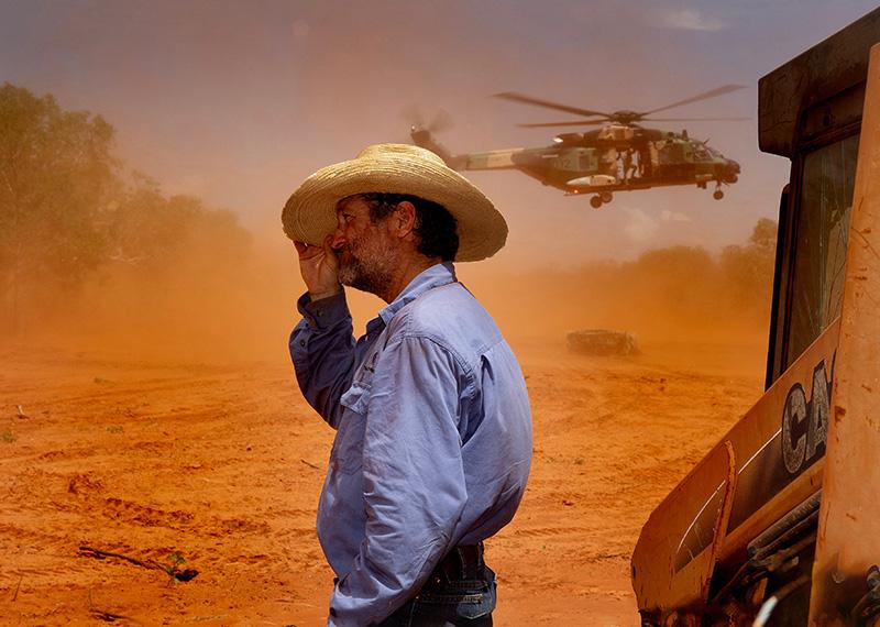 Man with cowboy hat standing on red dusty ground. There is also a military helicopter in the background. 